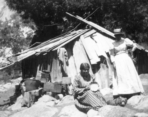 Two Native American (Miwok) women pose near a dilapidated wooden shack in Yosemite National Park, California. One woman, wearing a striped dress, sits holding a basket she is weaving. The other woman stands behind, wearing a long dress with leg o' mutton sleeves and a hat. The shack is loosely constructed and is surrounded by debris. A small bear is on the ground between the women.