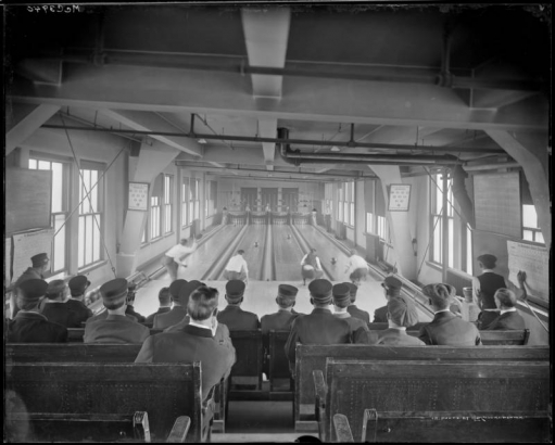Interior view of a bowling alley in Denver, Colorado; shows lanes, scorekeepers lettered: "Brunswick-Balke-Collender Co.," and rows of seated men wearing Denver Tramway Company conductor's hats.