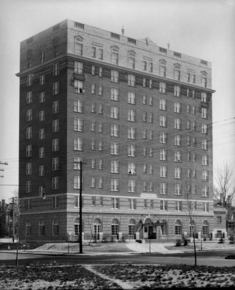 View of Colburn Hotel in Denver, Colorado; shows a brick building with urns and a covered entry. Cars are parked at the curb.