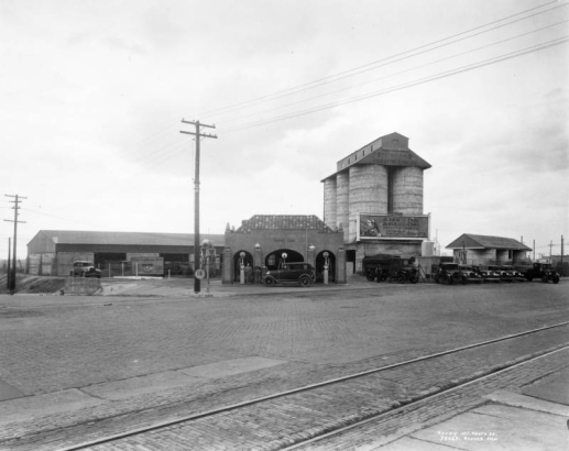 View of a coal dealership at 27th (Twenty-Seventh) and Walnut Streets, in Denver, Colorado; shows a stucco gasoline station with a tile roof, pumps, and signs: "The Colorado & Utah Coal Co.," "Harris Coal," "Drain Crankcase Here, Real Penn Motor Oils" and "White Gas." Men pose by cars, concrete silos are in the background, and streetcar tracks are by the sidewalk.