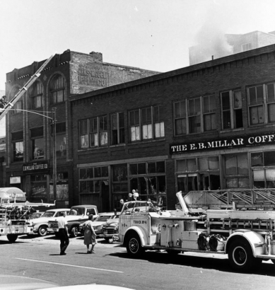 View of coffee dealers at 1822 Blake Street, during a fire, in Denver, Colorado; shows brick storefronts, cars, trucks, a fire truck, a fireman on a ladder, and signs: "The E. B. Millar Coffee Co."