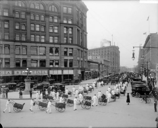 View of a parade at the intersection of Broadway and 16th (Sixteenth) Streets in Denver, Colorado; shows men in white uniforms with pushcarts, automobiles, the Majestic Building, the Brown Palace Hotel, and signs: "Colorado State & Savings Bank," "Miller Tires," and "The Texas Company."