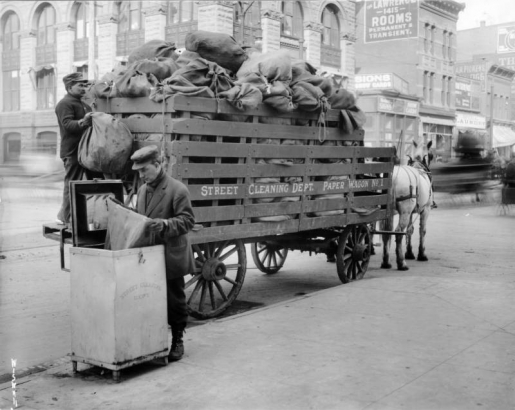 View of men working and a horse drawn-trash collection wagon on Arapahoe Street, in Denver, Colorado; brick and stone businesses are in the background. Lettering reads: "Street Cleaning Dept. Paper Wagon No. 1."