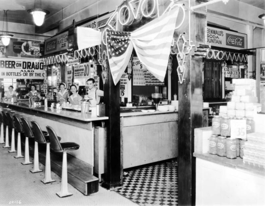 Interior view of a lunch counter at the Home Public Market, in Denver, Colorado; shows a man, women including Ruth Handler [third from right], a neon sign, tile floor, a United States flag, and signs: "Coca Cola," "Watermelon on Ice," "Beer On Draught," and "Greenwald's Soda Fountain."