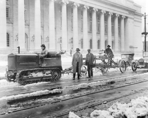View of Stout Street in Denver, Colorado; shows men working, a Caterpillar tractor, a snow plow, cars, and the United States Post Office.