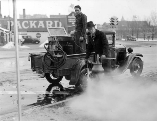 View of men working with a truck and spray-cleaning compressor, washing a fire hydrant in Denver, Colorado. A traffic signal is in the background.