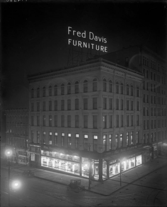 Nighttime rooftop view of Fred Davis Furniture Company at 15th (Fifteenth) and Larimer Streets in downtown Denver, Colorado. Automobiles are parked nearby. Signs read: "Complete Home Furnishers" and "Fred Davis Furniture Co."