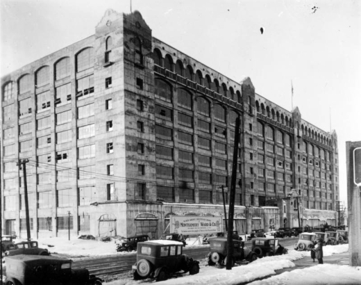 View of the newly constructed Montgomery Ward building at 501 South Broadway in the Baker neighborhood of Denver, Colorado. Automobiles and pedestrians are nearby. A sign near the building reads: "The New Rocky Mountain Home of Montgomery Ward & Co. The World's First Mail Order House Est. 1872 To Better Serve the People of Denver and the Mountain States."