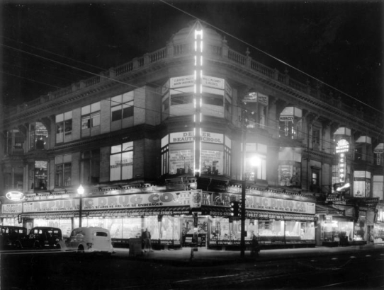 Nighttime view of the Enterprise Building on the corner of 15th (Fifteenth) and Champa Streets in downtown Denver, Colorado. Automobiles and pedestrians are near the building. Signs read: "Cut Rate Liquors and Wines," "The Purdy Studio," "Harco," "Geo. W. Fraser Tailor," "Katherine K. Corsets," "California Cotton Mills Co.," "Republic Drug Co.," "Denver Beauty School," "Coopers Flowers," and "Welcome V. F. W."
