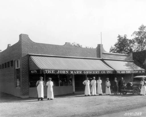 Employees pose outdoors near the John Marr Grocery Company in Denver, Colorado. The men wear aprons and caps. Lettering on the building's awnings read: "Mets," "The John Marr Grocery Co. Inc."