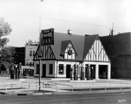 View of Frank L. Hansen's gas station at Broadway and Maple Avenue in the Baker neighborhood of Denver, Colorado. Fuel pumps and a two-car garage are near the building. A clock is above the main entryway. An electric sign reads: "Green Far-Go Gas."