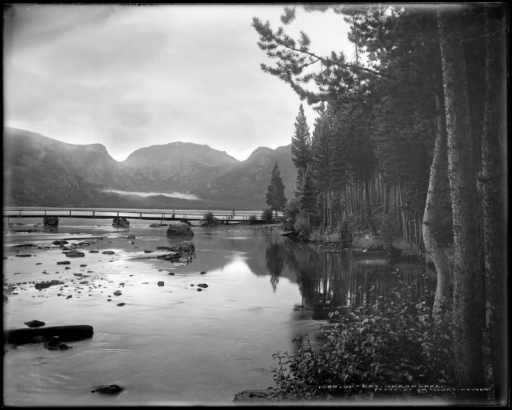 View of east inlet of Grand Lake, Colorado with Mt. Cairns, Mt. Craig, Mt. Adams, Mt. Wescott, Mt. Patterson and fog in the background. A footbridge spans the water; trees cast a reflection.