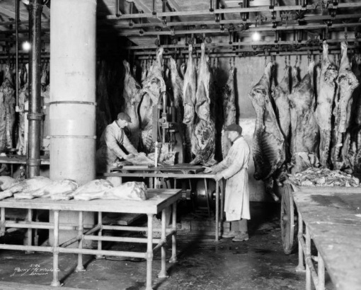 Interior view at Swift and Company in Denver, Colorado; shows men working, meat saws, and hanging sides of beef.