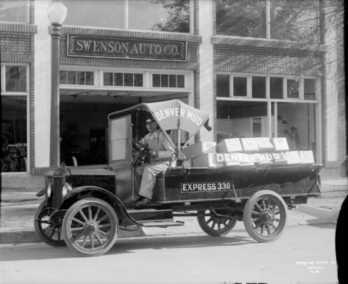 A man sits in a delivery truck parked near the Swenson Auto Company at 1960 Champa Street in downtown Denver, Colorado. Lettering on the truck reads: "Denver Mud Express 330."