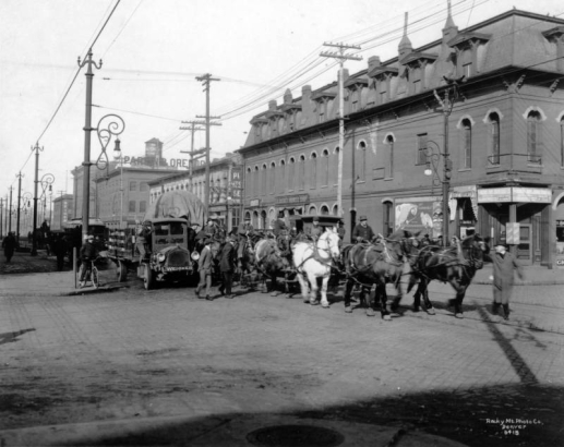 A team of horses pull a wagon with a large canvas covered object on Blake Street at 15th (Fifteenth) Street in downtown Denver, Colorado. Men walk nearby. Signs on buildings read: "The N. J. O'Fallon Supply Co.," "Goldberg's Harness Shop," "Soft Drinks," and "Cigars." A pick-up truck is beside the wagon.