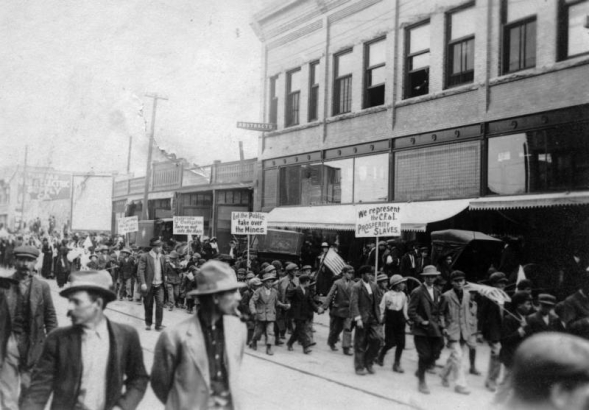 Boys march in support of UMW coal miners on strike against CF&I in Trinidad, Las Animas County, Colorado, carrying signs: "We Represent the CF&I Prosperity Slaves," "Let The Public take Over The Mines,"  "Colorado Civilization Turn Us Out in the Rain," and "Has the Governor any Respect for the State?" Men crowd sidewalks by brick businesses.