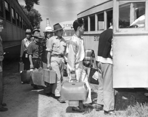 Japanese men arrive from the Merced Assembly Center, California, by train, carrying suitcases and boarding school buses on their way to Camp Amache, Granada Relocation Center, Prowers County, southeastern Colorado. A United States soldier in uniform stands next to the passenger train car and looks on as the line of evacuees board the bus.