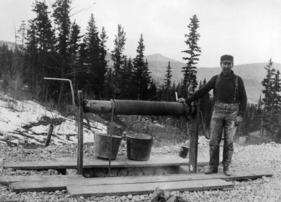 A man stands near a crank well hoist. Two buckets are attached to the rope.