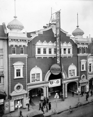 Rooftop view of the Empress Theater at 1615 Curtis Street in downtown Denver, Colorado. Pedestrians walk nearby. The three-story building has decorative trim and is topped with minarets. Signs on the building read: "Pantage's Vaudville" and "Now Showing 10 Acts Spring Festival Show."