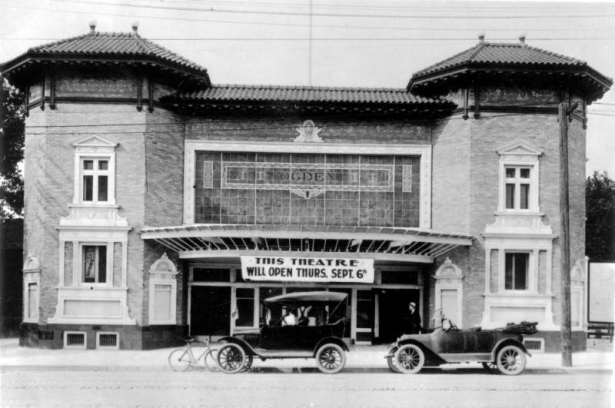 View of the Ogden Theater on Colfax Avenue in Denver, Colorado. The two-story brick building has a tile roof and ornate entryway. Automobiles and a bicycle are parked nearby. A sign reads: "This Theater Will Open Thurs. Sept. 6th."
