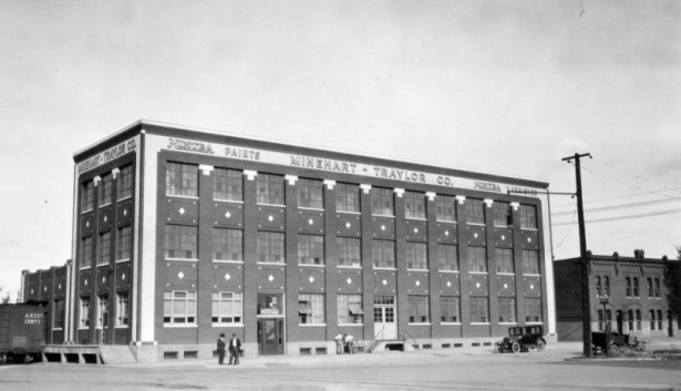 Men stand near the Minehart-Traylor Company building at the intersection of 25th (Twenty-fifth) Street, Broadway, and Walnut Street in the Five Points neighborhood of Denver, Colorado. The three-story brick building has decorative brickwork and contrasting trim. Lettering on the building reads: "Mintra Paints Minehart-Traylor Co."
