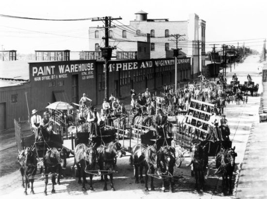 Employees of the McPhee-McGinnity Manufacturing Company pose on horse-drawn wagons near the company's warehouse at 23rd (Twenty-third) and Blake Streets in the Five Points neighborhood of Denver, Colorado. Lettering on the one-story corrugated metal building reads: "Paint Warehouse McPhee and McGinnity Company."