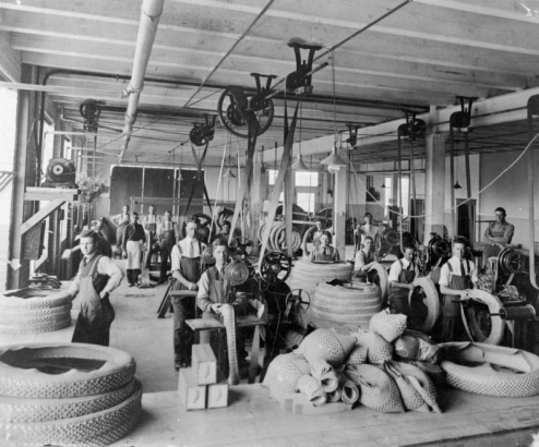 Interior view of the Gates Rubber Company in Denver, Colorado; shows men working with belt-driven machinery and rubber tires.
