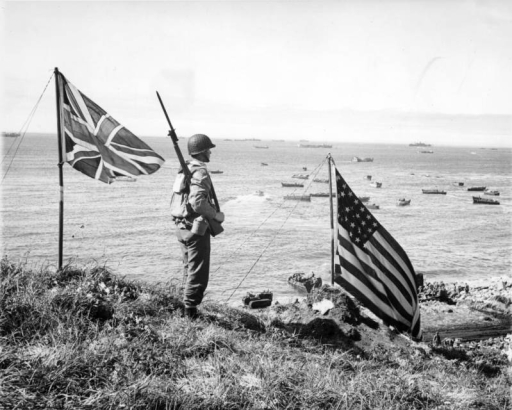 Jim "Wyoming" Penton, a member of the 10th Mountain Division 87th Regiment Service Company (SVC), stands on a bluff over Broad Beach on Kiska Island in Alaska; U.S. and British flags are nearby. The 87th Mountain Infantry Regiment was in the Aleutians as a part of Amphibious Task Force 9. Boats and amphibious vehicles are in the water in the distance.