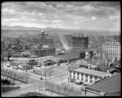 View of Denver, Colorado, shows downtown buildings including the Arapahoe County Courthouse. Clouds top front range mountains; signs read: "I.A. (Mrs. Ida Alkire) Lehman Specialty Shop (or The Toggler Shop, 1538 California)", "Overland Cars," and "YMCA."