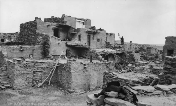 A Native American (Hopi) woman stands near a group of adobe cluster homes at Oraibi Pueblo, Arizona. The homes have chimney pots and wooden vigas.