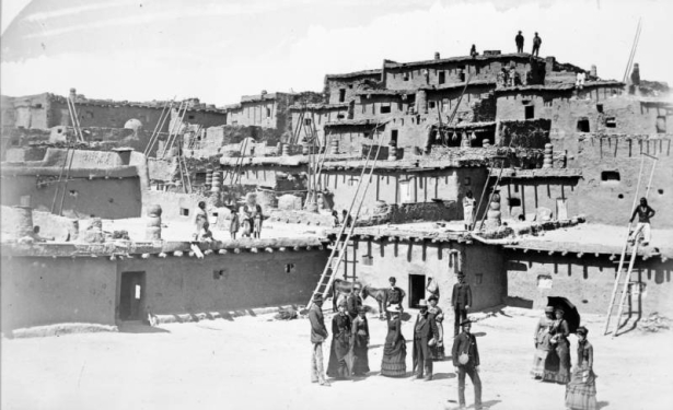 Native American (Pueblo, Zuni) children and women and white men and women stand on and near adobe and stone cluster homes at Zuni Pueblo, New Mexico. The buildings have chimney pots, vigas, and wooden ladders.