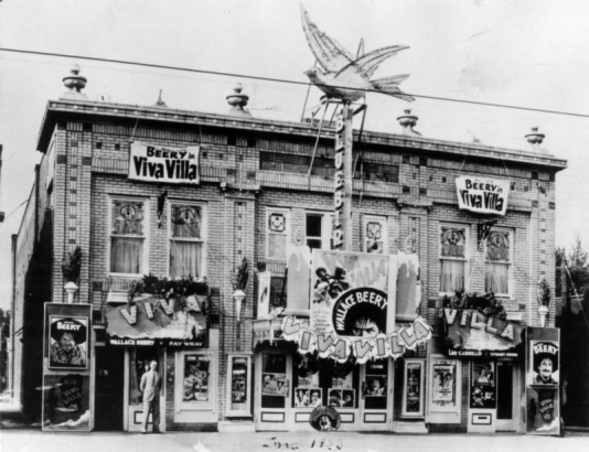 View of the Bluebird Theater at 3317 East Colfax Street in Denver, Colorado; electric sign depicts a bird and reads: "Bluebird," banners read: "Wallace Beery in Viva Villa." Corbeled brick and urns ornament the building.