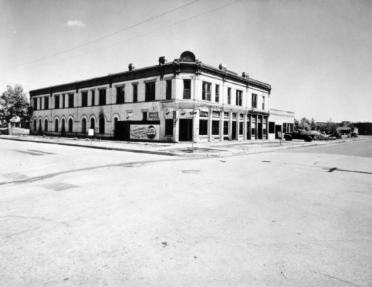 View of a building on the corner of 26th and Arapahoe Streets in the Five Points neighborhood of Denver, Colorado. The two-story building has display windows and arched window wells. Signs read: "More Bounce to the Ounce Pepsi-Cola" and "Coca-Cola."