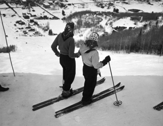 Two women skiers in Aspen, Colorado stand at the top of the ski hill facing downwards. Both women wear trousers, light jackets and scarves on their head.  Trees are on either side of the ski slope, and the town is at the base of the hill.