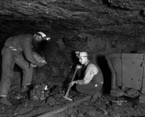 View of two miners working in an underground coal mine, Lafayette, Colorado. One man squats and uses his pickaxe on a chunk of coal, the other holds an unidentified piece of equipment. An empty ore cart is next to them.