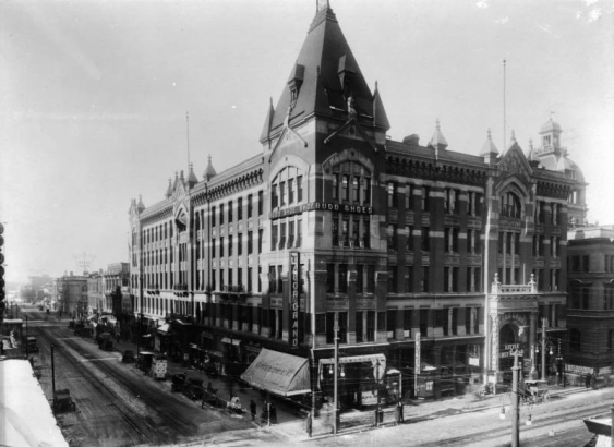 View of the Tabor theater building at 16th (Sixteenth) and Curtis Streets in Denver, Colorado. Horse-drawn merchants wagons and cars are in the street; signs read: "Budd Shoes," "Tabor Grand," "Little Lost Sister," and "Joseph Schwartz Rare Gems."