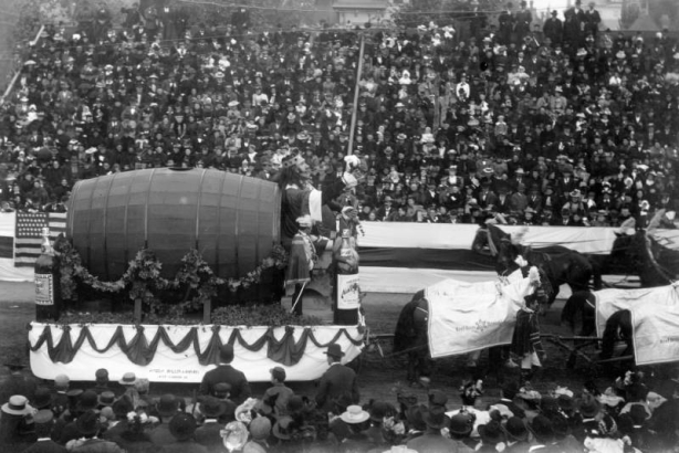 Men ride on a horse-drawn float, part of the Festival of Mountain and Plain, in a parade in Denver, Colorado. The float depicts a king with a sword and a keg of beer. Men on the float wear costumes that include hats decorated with feathers, capes, and swords. The horses wear decorative cloth shrouds with lettering that reads: "Neef Bros Brewing Co." Spectators sit in grandstands nearby.