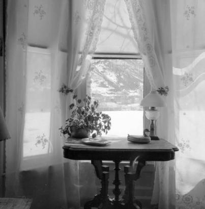 Interior view of a rectangular table by a bay window in the home of Fred Glidden (Luke Short) in Aspen, Colorado; features a kerosene lamp with a glass globe, duck sculpture, and ashtray on the table; sheer curtains with embroidered floral patterns cover the windows; mountain covered with snow in the background.