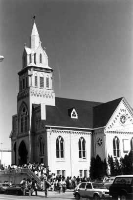 Hispanic-American men, women, and children leave after Easter Mass at the Sacred Heart Catholic Church at 2760 Larimer Street in the Five Points (Curtis Park) neighborhood of Denver, Colorado. The church building has a bell tower, stained glass windows, decorative lintels, and an arched entryway.