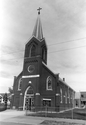 View of Saint Joseph Polish Church at 517 East 46th (Forty-sixth) Avenue in the Globeville neighborhood of Denver, Colorado. The brick building has a bell tower, spire, and circular and arched stained glass windows. A banner reads (in Polish): "Kosciol Sw Jozefa."
