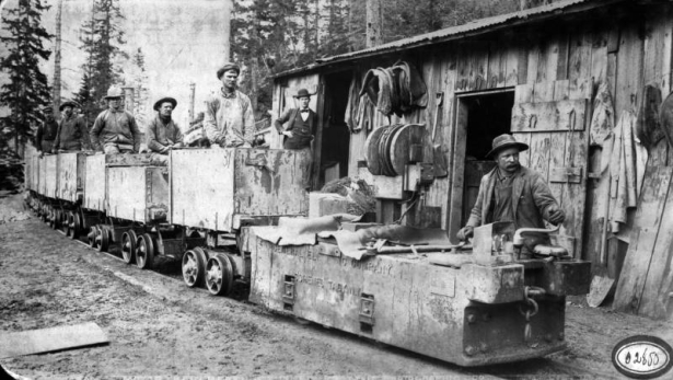 View of men in chain of ore carts being pulled by General Electric engine with frame building, a man stands beside the door, Colorado.