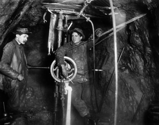 Two miners working by candlelight tend a diamond drill rig one mile underground in the Camp Bird mine, Ouray County, Colorado.