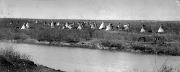 Panoramic view of a Native American (Kiowa or Apache) tepee camp by a river near Fort Sill, Indian Territory. Wagons are parked in the camp.