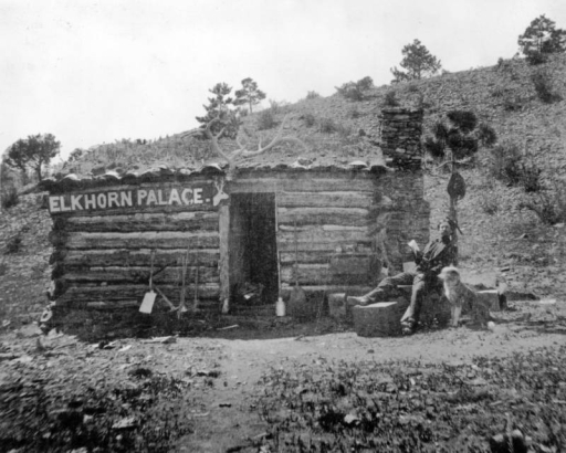 A man sits with his dog outside of a hewn log cabin near Rosita (Custer County), Colorado. The cabin has a single doorway, a rack of elk horns over the door, a chimney, and a sign reading: "Elkhorn Palace."