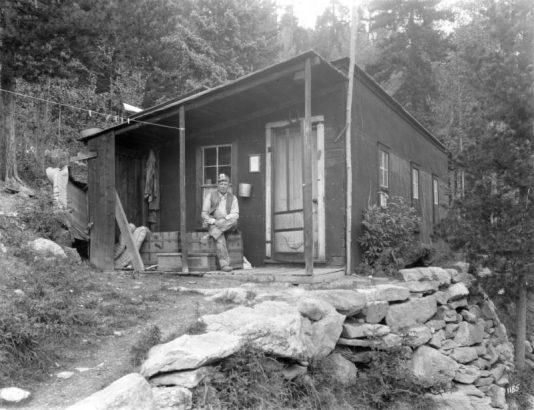 A miner poses outdoors on a trunk on the porch of his cabin probably in Colorado. He wears a cap with a miner's light.