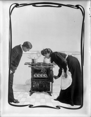 A photomontage of a couple as they look into the open oven of a gas stove. Steam rises from a pot on the stove and from food in the oven. The woman wears a dark dress and white apron; the man wears a suit and white shirt. The image is surrounded with a decorative border.