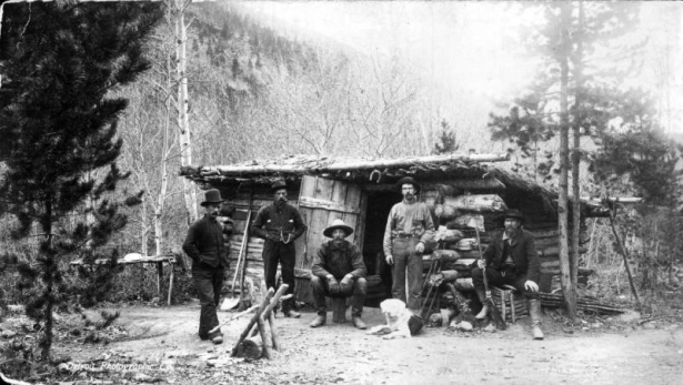View of five miners and dog at log cabin with dirt roof, Colorado.