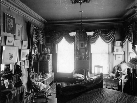 Interior view of the Bancroft house in Denver, Colorado; decor includes a bed, wicker rocking chair, dresser, pillows, and framed art including a Winslow Homer watercolor.