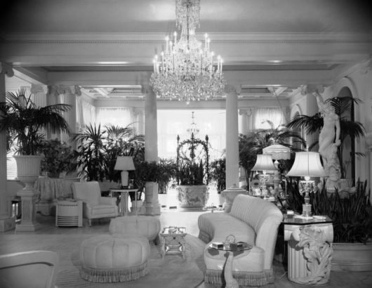 Interior view of the Claude K. Boettcher residence, in Denver, Colorado; decor includes upholstered couches, chairs, ottomans, palms, a sculpture of a woman, lamps, a crystal chandelier, classical columns, and a planter with bas-relief ornaments.