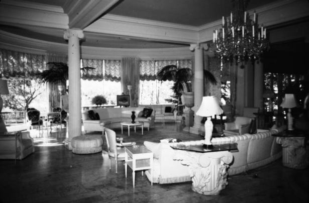 Interior view at the Claude K. Boettcher residence, in Denver, Colorado; living room decor includes curved couches, chairs, an ottoman, glass tables, Corinthian capitols, a crystal chandelier, palms, and curtains.